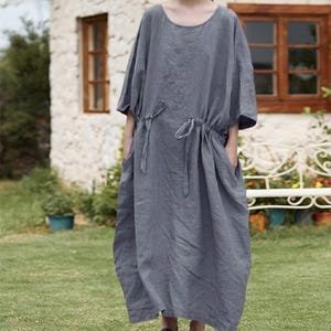 May include: A gray linen dress with a round neckline and short sleeves. The dress features a drawstring waist and pockets. The model is wearing dark brown shoes. The dress is suitable for casual wear.