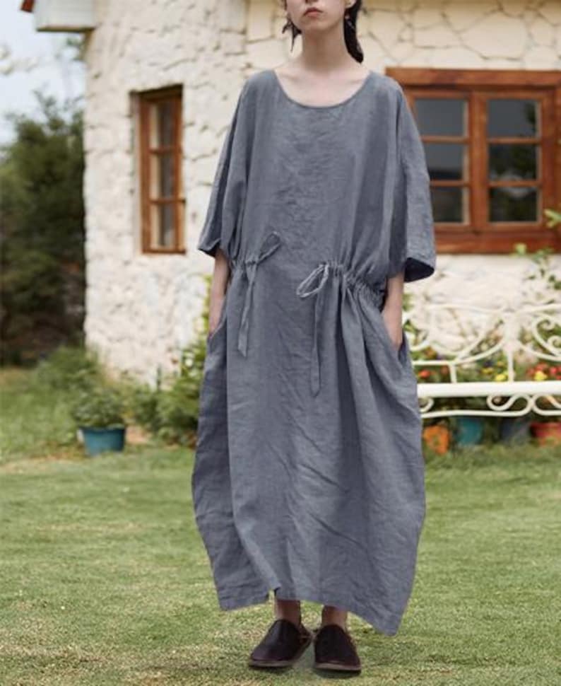 May include: A gray linen dress with a round neckline and short sleeves. The dress features a drawstring waist and pockets. The model is wearing dark brown shoes. The dress is suitable for casual wear.