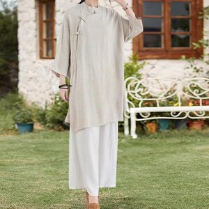 May include: A beige linen tunic top with a mandarin collar and asymmetrical hemline, paired with white wide-leg pants. The outfit is completed with brown shoes. The ensemble is suitable for casual wear.