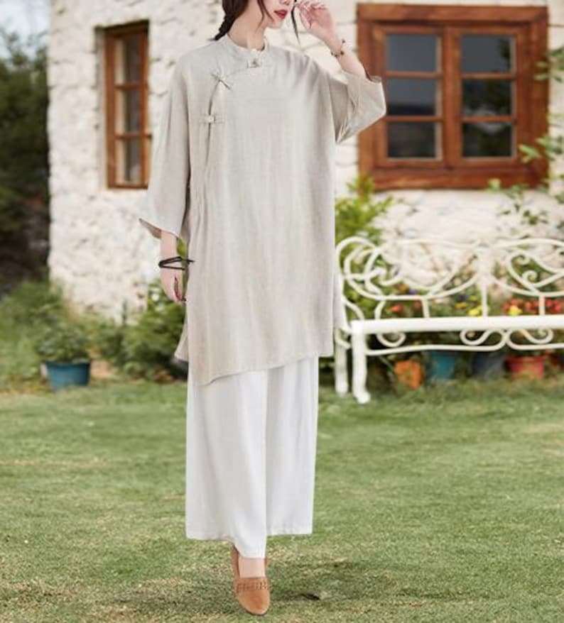 May include: A beige linen tunic top with a mandarin collar and asymmetrical hemline, paired with white wide-leg pants. The outfit is completed with brown shoes. The ensemble is suitable for casual wear.