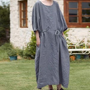May include: A long, gray linen dress with a round neckline and short, wide sleeves. The dress features a drawstring waist and pockets. The model is wearing brown leather shoes. The dress is suitable for casual wear.