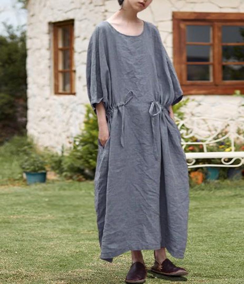 May include: A long, gray linen dress with a round neckline and short, wide sleeves. The dress features a drawstring waist and pockets. The model is wearing brown leather shoes. The dress is suitable for casual wear.
