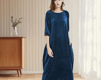 May include: A long, dark blue velvet dress with a round neckline and three-quarter sleeves. The dress has pockets and a loose, flowing silhouette. The model is wearing tan shoes. The background includes a wooden cabinet and a white curtain.
