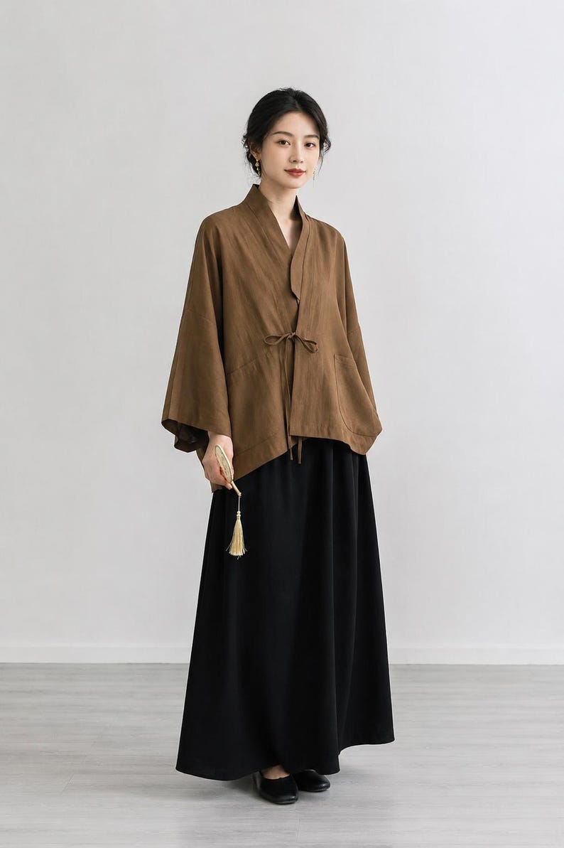 May include: A brown wrap-style jacket with wide sleeves and a thin tie closure at the waist, paired with a long, solid black skirt. The jacket features a V-neckline and a structured collar, while the skirt falls to the ankles in a simple, straight silhouette.