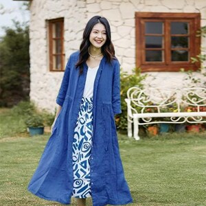 May include: A woman wearing a long, cobalt blue linen duster coat over a white top and a patterned skirt. The skirt features a white background with blue abstract shapes. The coat is buttoned down the front and has three-quarter sleeves.