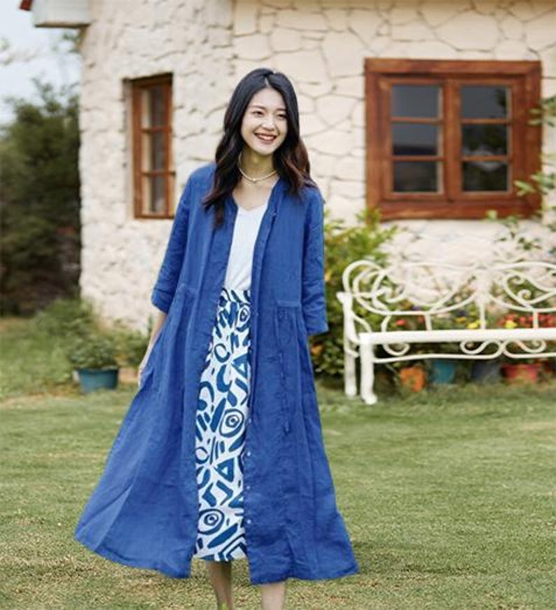 May include: A woman wearing a long, cobalt blue linen duster coat over a white top and a patterned skirt. The skirt features a white background with blue abstract shapes. The coat is buttoned down the front and has three-quarter sleeves.