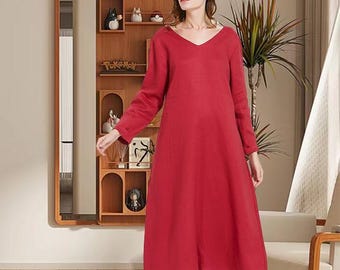 May include: A long, red dress with a V-neck and long sleeves. The dress is knee-length and appears to be made of a soft, flowing fabric. The dress is worn by a person in a room with a wooden shelf and a plant.