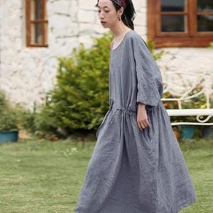 May include: A woman wearing a long, gray linen dress with a drawstring waist and pockets. She is wearing brown leather shoes and a patterned headband. The background includes a stone wall, wooden window frames, and green grass.