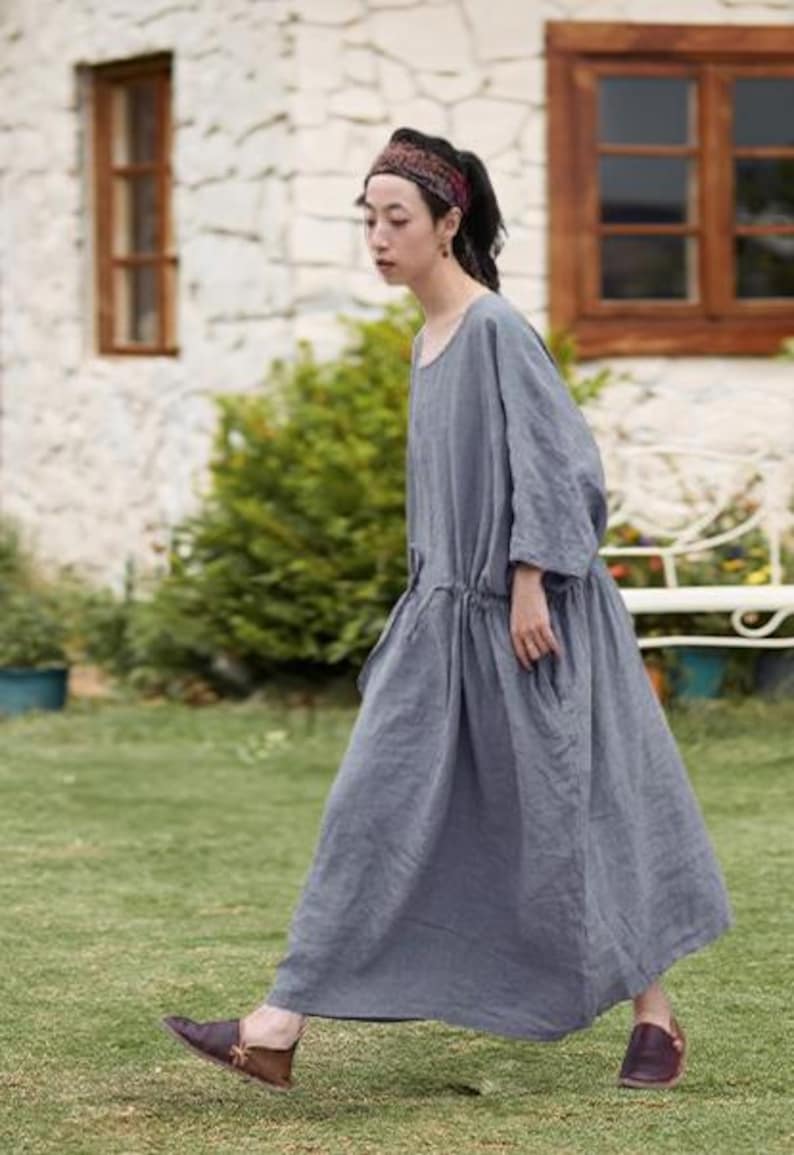 May include: A woman wearing a long, gray linen dress with a drawstring waist and pockets. She is wearing brown leather shoes and a patterned headband. The background includes a stone wall, wooden window frames, and green grass.