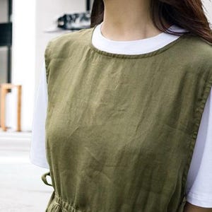 May include: An olive green linen apron dress over a white t-shirt. The dress has a round neckline, a fitted waist, and a flowing skirt. The fabric appears to be lightweight and breathable, suitable for warm weather. The overall style is casual and minimalist.