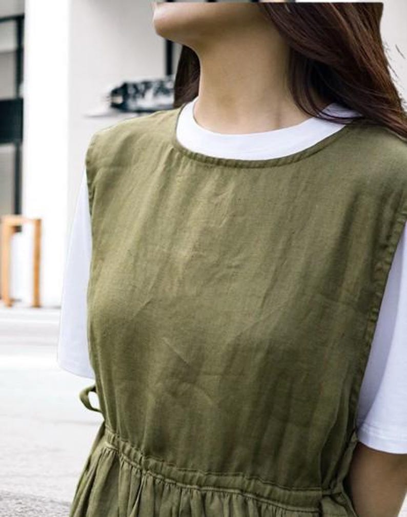 May include: An olive green linen apron dress over a white t-shirt. The dress has a round neckline, a fitted waist, and a flowing skirt. The fabric appears to be lightweight and breathable, suitable for warm weather. The overall style is casual and minimalist.