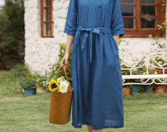 May include: A blue linen dress with a square neckline and a tie belt. The dress has three-quarter sleeves and buttons down the front. A woven basket filled with sunflowers and other flowers is held in the hand.