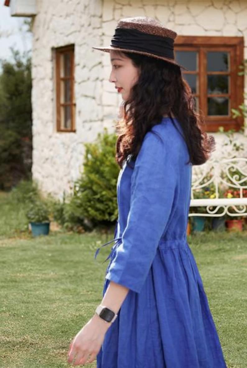 May include: A woman wearing a blue dress and a straw hat with a black band. The dress has three-quarter sleeves and a drawstring waist. The hat has a wide brim. The background includes a white stone building and green grass.
