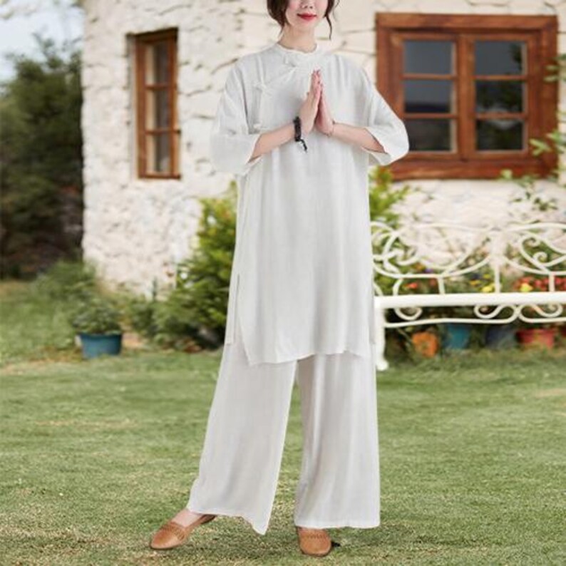 May include: A white linen tunic and wide-leg pants set. The tunic has a mandarin collar and side slits. The pants are loose-fitting. The outfit is worn with brown flats. The background includes a white building, green grass, and a white bench.