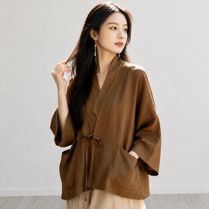 May include: A brown wrap-style jacket with three-quarter length sleeves, a V-neckline, and a thin tie closure at the waist. The garment features two large patch pockets on the front and is paired with a light beige, layered, flowing skirt.