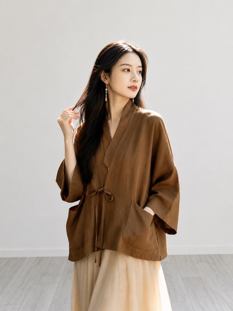 May include: A brown wrap-style jacket with three-quarter length sleeves, a V-neckline, and a thin tie closure at the waist. The garment features two large patch pockets on the front and is paired with a light beige, layered, flowing skirt.