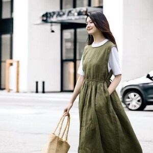 May include: An olive green linen dress with a white t-shirt underneath. The dress has a gathered waist and pockets. A woman is carrying a woven tote bag with long handles. The background includes a building and a car.