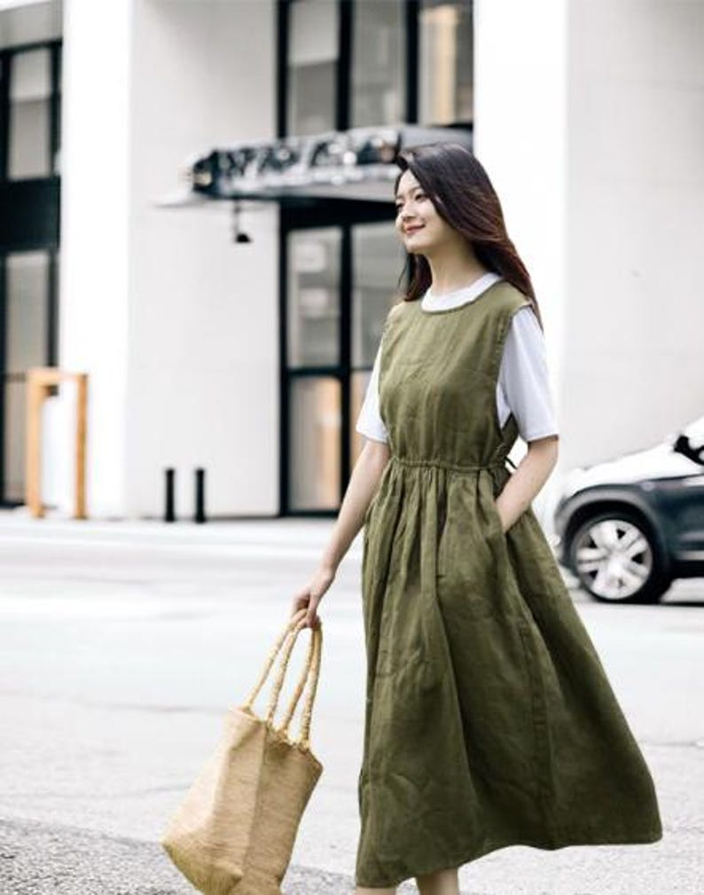 May include: An olive green linen dress with a white t-shirt underneath. The dress has a gathered waist and pockets. A woman is carrying a woven tote bag with long handles. The background includes a building and a car.