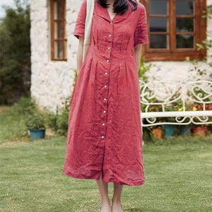 May include: A red midi dress with short sleeves and a button-down front. The dress has a collared neckline and pleats at the waist. The model is wearing white flats and carrying a cream-colored tote bag. The background includes a white building and green grass.