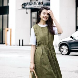 May include: An olive green linen dress with a white t-shirt underneath. The dress has a fitted bodice and a flowing skirt. A woven straw tote bag is held in the hand. The background includes a building and a car.