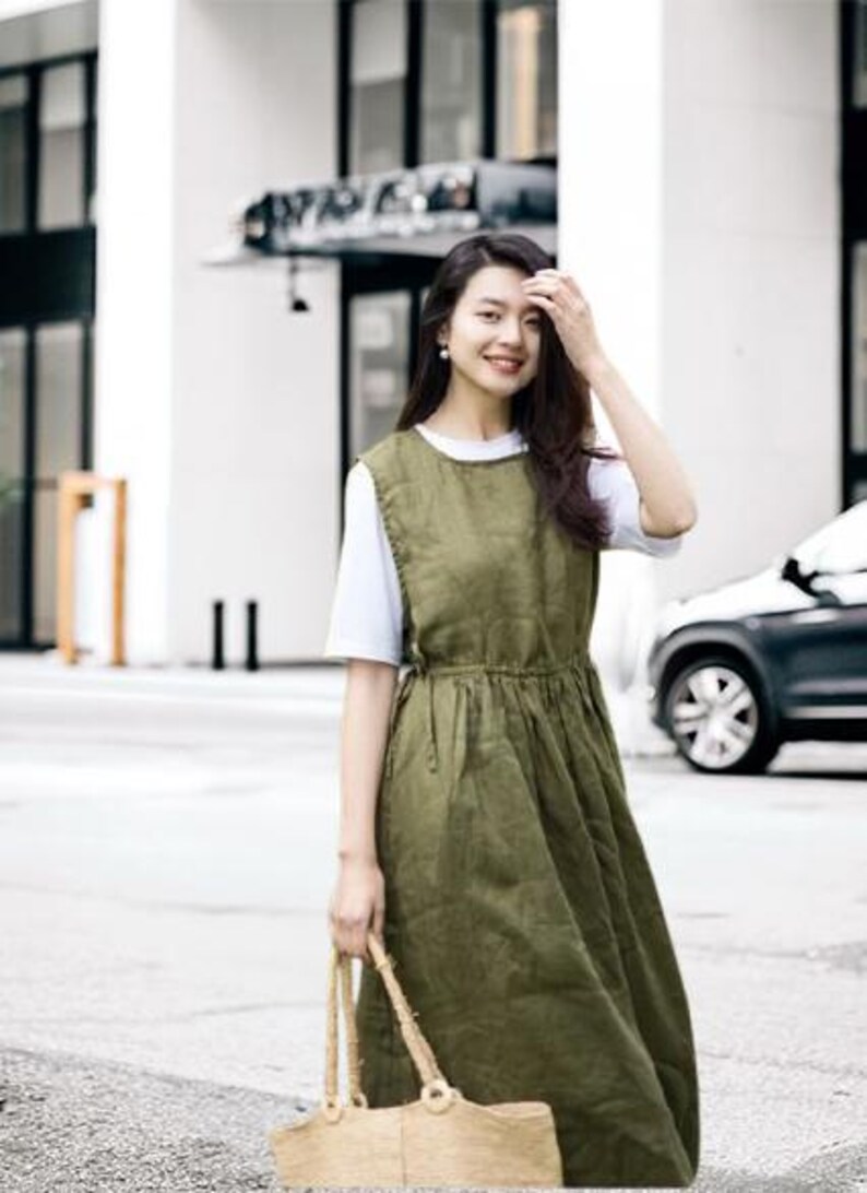 May include: An olive green linen dress with a white t-shirt underneath. The dress has a fitted bodice and a flowing skirt. A woven straw tote bag is held in the hand. The background includes a building and a car.