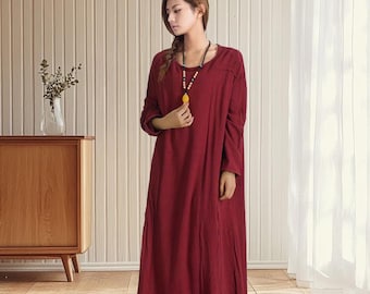 May include: A long, burgundy-colored dress with long sleeves. The dress has a simple, flowing design and is worn with a necklace. The model is also wearing light-colored shoes. The dress is made of a soft, flowing fabric.