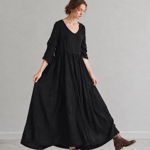 May include: A floor-length black dress with a V-neckline, three-quarter length sleeves, and a gathered waistline that creates a full, flowing skirt. The garment is paired with brown leather ankle boots, completing the overall ensemble.