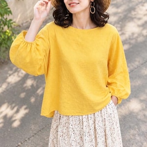 May include: A bright yellow long-sleeved blouse with a round neckline and gathered cuffs, paired with a white skirt featuring a small black and brown speckled pattern. A woven straw hat with a wide brim is worn on the head.