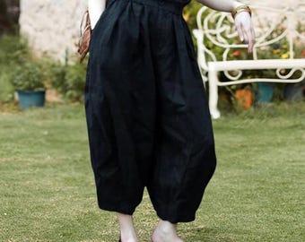 Puede incluir: Pantalones negros anchos y cortos, de cintura alta. Los pantalones están hechos de una tela suave y tienen un corte holgado. La modelo lleva sandalias marrones y una blusa azul. El fondo es un césped verde con un banco blanco.
