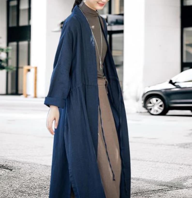 May include: A long, navy blue linen coat with a drawstring waist and long sleeves. The coat is worn over a brown top and skirt. A silver necklace is visible. The outfit is suitable for layering and offers a relaxed, yet stylish look.