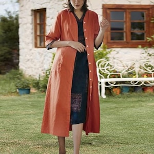 May include: A long, button-down, rust-colored linen duster coat over a dark blue patterned dress. The coat has three-quarter sleeves with dark blue trim. The dress has a V-neck and a patterned panel at the hem. The outfit is worn outdoors.