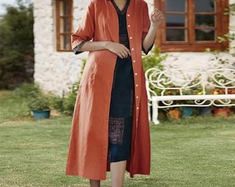May include: A long, button-down, rust-colored linen duster coat over a dark blue patterned dress. The coat has three-quarter sleeves with dark blue trim. The dress has a V-neck and a patterned panel at the hem. The outfit is worn outdoors.
