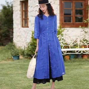 May include: A woman wearing a cobalt blue linen dress with a button-down front, three-quarter sleeves, and a tie waist. She is also wearing a straw hat with a dark band, blue sandals, and carrying a white tote bag. The dress falls to the mid-calf.