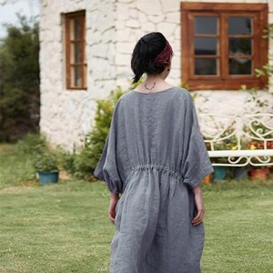May include: A person wearing a gray linen dress with a drawstring waist, standing in a grassy yard. The dress has short sleeves and a loose fit. A burgundy headband is worn. A stone house with brown window frames is in the background.