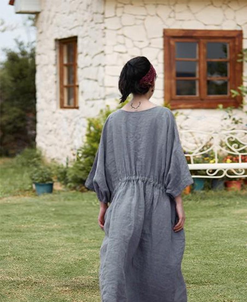 May include: A person wearing a gray linen dress with a drawstring waist, standing in a grassy yard. The dress has short sleeves and a loose fit. A burgundy headband is worn. A stone house with brown window frames is in the background.