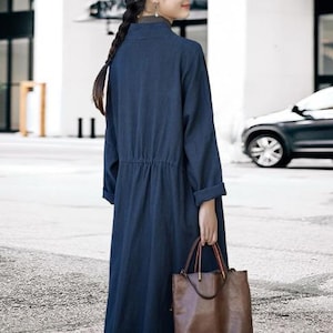 May include: A long, navy blue dress with long sleeves and a cinched waist. The dress is paired with a brown leather tote bag with red accents. The dress has a mandarin collar and is made of a soft, flowing fabric.