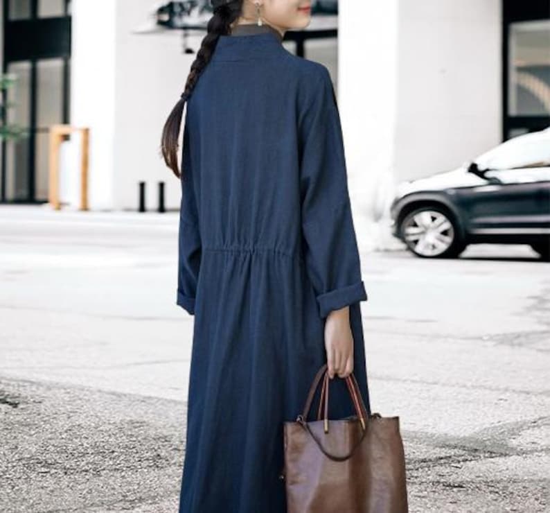 May include: A long, navy blue dress with long sleeves and a cinched waist. The dress is paired with a brown leather tote bag with red accents. The dress has a mandarin collar and is made of a soft, flowing fabric.