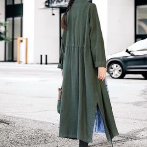 May include: A long, olive green coat with a drawstring waist and side slits. The coat has long sleeves and a mandarin collar. The person is wearing black leggings and brown boots. A woven bag is carried in the left hand.