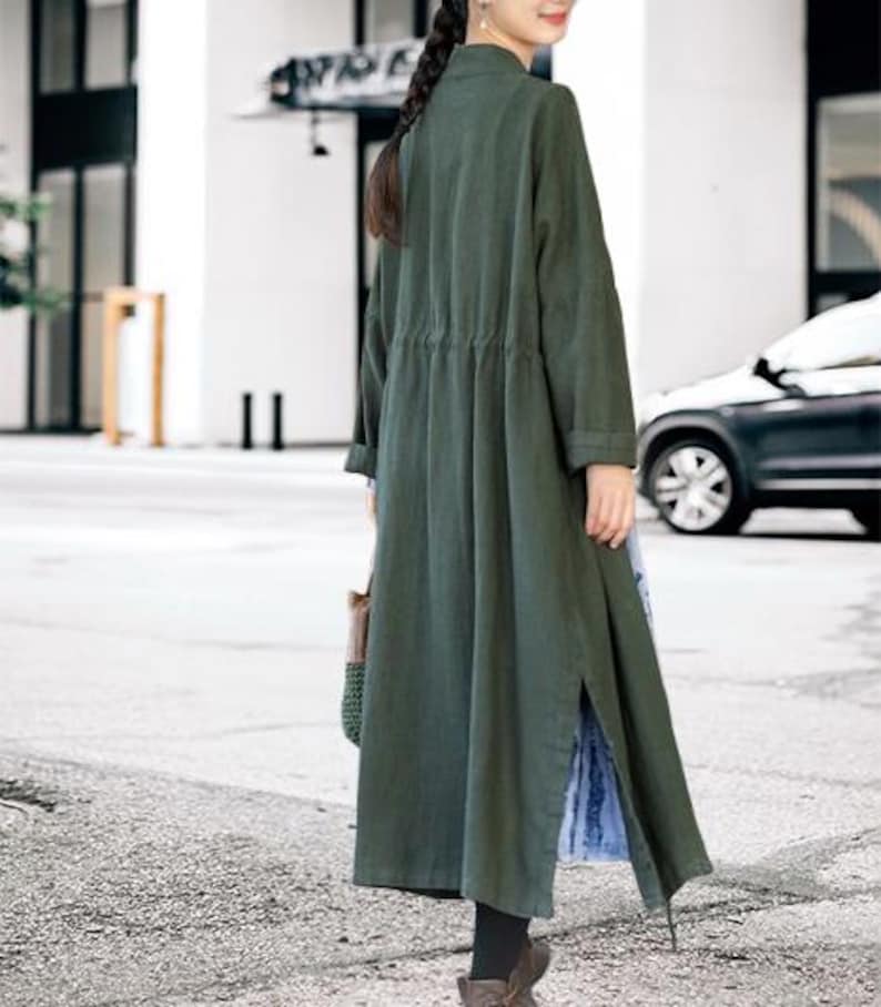 May include: A long, olive green coat with a drawstring waist and side slits. The coat has long sleeves and a mandarin collar. The person is wearing black leggings and brown boots. A woven bag is carried in the left hand.