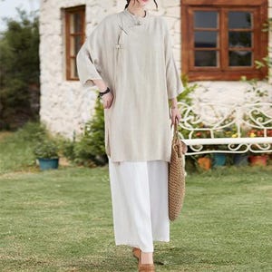 May include: A beige linen tunic with a mandarin collar and side slits, paired with white wide-leg pants. The outfit is accessorized with a woven tote bag and brown sandals. The ensemble is suitable for casual wear.