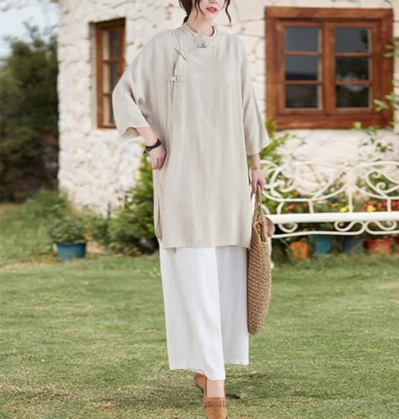 May include: A beige linen tunic with a mandarin collar and side slits, paired with white wide-leg pants. The outfit is accessorized with a woven tote bag and brown sandals. The ensemble is suitable for casual wear.