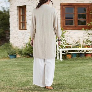 May include: A beige linen duster coat with three-quarter sleeves and white wide-leg pants. The outfit is worn outdoors on a grassy lawn. The duster coat is knee-length and has a relaxed fit. The pants are loose and flowy.