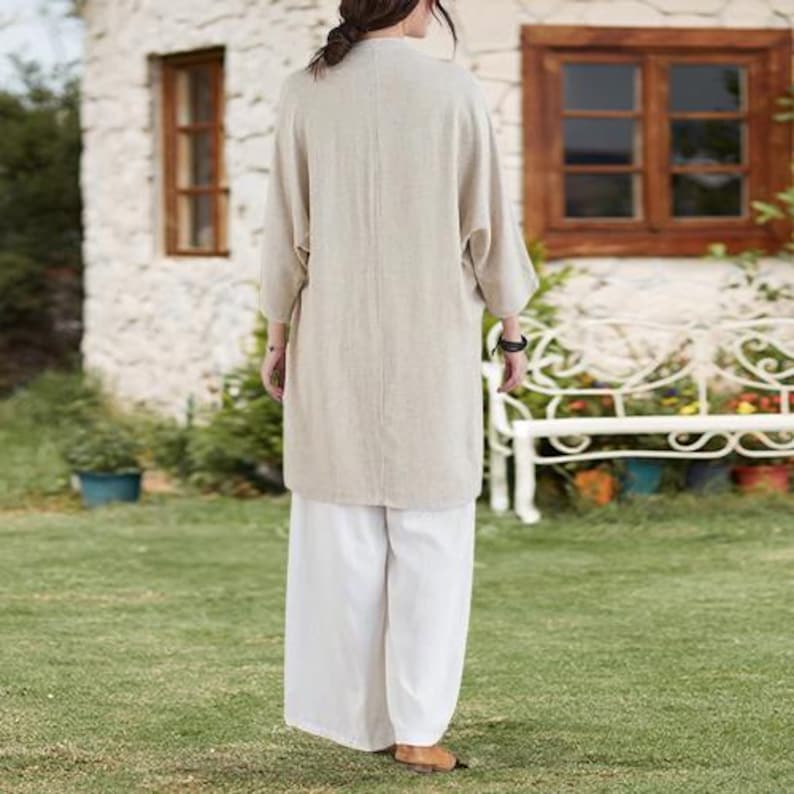 May include: A beige linen duster coat with three-quarter sleeves and white wide-leg pants. The outfit is worn outdoors on a grassy lawn. The duster coat is knee-length and has a relaxed fit. The pants are loose and flowy.
