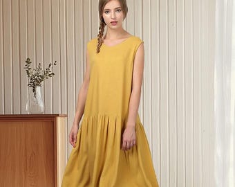 Peut inclure: Robe sans manches jaune moutarde avec une taille froncée et une jupe fluide. La robe arrive au genou et est portée avec des chaussures en cuir marron. En arrière-plan, un meuble en bois et un mur texturé.