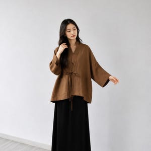 May include: A brown long-sleeved jacket with a V-neckline and a thin tie closure at the waist, paired with a solid black ankle-length skirt and black flat shoes. The jacket features wide sleeves and a loose, relaxed fit throughout the garment.