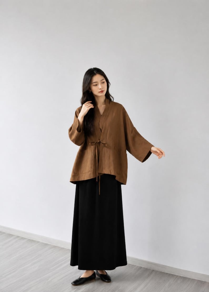 May include: A brown long-sleeved jacket with a V-neckline and a thin tie closure at the waist, paired with a solid black ankle-length skirt and black flat shoes. The jacket features wide sleeves and a loose, relaxed fit throughout the garment.