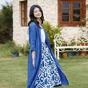 May include: A woman wearing a blue linen duster over a white top and a patterned skirt. The skirt features a white background with blue abstract shapes. The outfit is worn outdoors in front of a stone building and a white bench.
