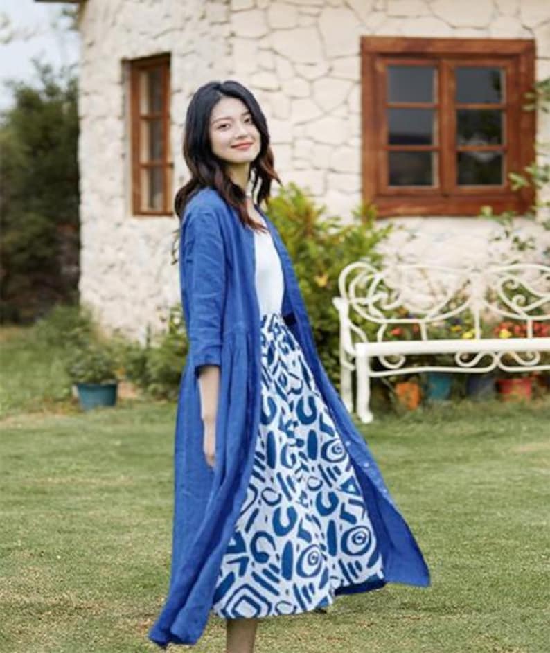 May include: A woman wearing a blue linen duster over a white top and a patterned skirt. The skirt features a white background with blue abstract shapes. The outfit is worn outdoors in front of a stone building and a white bench.