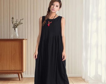 May include: A sleeveless black dress with a gathered waist and midi length. The dress is worn with a red pendant necklace and tan sandals. The background features a wooden cabinet, vertical wood paneling, and a sheer white curtain.