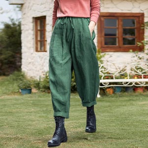 Könnte beinhalten: Grüne Leinenhose mit Kordelzug und Taschen. Die Hose wird mit schwarzen Kampfstiefeln kombiniert. Die Person trägt ein langärmliges rosa Oberteil. Das Outfit wird vor dem Hintergrund eines Steingebäudes und grünen Grases präsentiert.